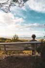 Вид туриста в кепке, сидящего на деревянной скамейке под деревом и смотрящего на пейзаж под облачным небом — стоковое фото