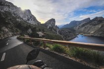 Зверху водій врожаю їде на мотоциклі по дорозі в оточенні гір біля озера проти хмарного неба — стокове фото