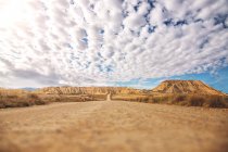 Leere Straße mit brauner Erde geht in die Ferne zwischen trockenen Sträuchern und braunen Bergen und blauem Himmel mit weißen Wolken auf dem Hintergrund bei Bardenas Reales — Stockfoto