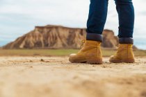 Legs on faceless traveler in brown boots and blue jeans standing on dirty sandy road with mountain and sky on blurred background in Bardenas Reales, Navarre, Spain — Stock Photo