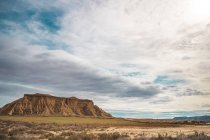 Champ vide avec végétation clairsemée verte et falaise brune puissante en arrière-plan sous un ciel bleu nuageux à Bardenas Reales, Navarre, Espagne — Photo de stock