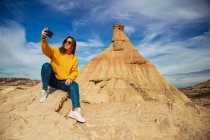 Радісна молода жінка - мандрівниця в стильному повсякденному одязі посміхається, коли робить це на мобільному телефоні з коричневим пагорбом і синім небом на задньому плані в Барденас - Реалес (Наварра, Іспанія). — стокове фото