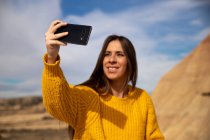 Fröhliche junge Reisende in stylischer Freizeitkleidung lächeln, während sie ein Selfie auf dem Handy mit braunem Hügel und blauem Himmel im Hintergrund in Bardenas Reales, Navarra, Spanien machen — Stockfoto