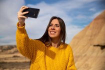 Joyful young female traveler in stylish casual wear smiling while taking selfie on mobile phone with brown hill and blue sky on background in Bardenas Reales, Navarre, Spain — Stock Photo