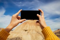 Обрезать женские руки анонимного путешественника фотографируя большой коричневый скала с красочным голубым небом и белые облака на заднем плане в Барденас Реалес в Испании — стоковое фото
