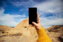 Recorte manos femeninas de viajero anónimo tomando fotos de un gran acantilado marrón con cielo azul colorido y nubes blancas en el fondo de Bardenas Reales en España - foto de stock