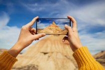 Cultivez les mains féminines d'un voyageur anonyme prenant une photo d'une grande falaise brune avec un ciel bleu coloré et des nuages blancs en arrière-plan à Bardenas Reales en Espagne — Photo de stock