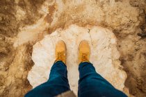 From above top view of anonymous person legs in brown boots and blue jeans standing on dirty sandy road with mountain and sky on blurred background in Bardenas Reales, Navarre, Spain — Stock Photo