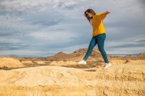 Vista lateral da jovem turista em camisola amarela jeans azul e tênis branco espalhando braços para equilíbrio enquanto caminha ao longo do pico da colina marrom em Bardenas Reales, Navarra, Espanha — Fotografia de Stock
