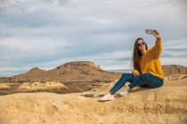 Радостная молодая женщина в стильной повседневной одежде улыбается, делая селфи на мобильном телефоне на фоне коричневого холма и голубого неба в Барденас-Реалес, Наварра, Испания — стоковое фото
