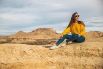 Joyeuse jeune voyageuse en tenue décontractée élégante assise dans une colline brune avec un ciel bleu en arrière-plan à Bardenas Reales, Navarre, Espagne — Photo de stock
