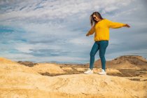 Vista lateral da jovem turista em camisola amarela jeans azul e tênis branco espalhando braços para equilíbrio enquanto caminha ao longo do pico da colina marrom em Bardenas Reales, Navarra, Espanha — Fotografia de Stock