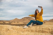 Joyful jovem viajante em elegante casual desgaste sorrindo ao tirar selfie no telefone móvel com colina marrom e céu azul no fundo em Bardenas Reales, Navarra, Espanha — Fotografia de Stock