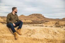Low angle of content bearded male in casual wear and sunglasses smiling while resting at foot of brown mountain with blue sky on background at Bardenas Reales — Stock Photo