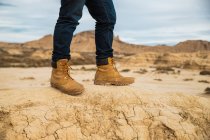 Ноги безликого путешественника в коричневых сапогах и синих джинсах стоят на грязной песчаной дороге с горами и небом на размытом фоне в Барденас Реалес, Наварра, Испания — стоковое фото