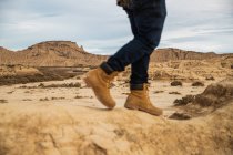 Jambes sur un voyageur sans visage en bottes brunes et jeans bleus debout sur une route sablonneuse sale avec montagne et ciel sur fond flou à Bardenas Reales, Navarre, Espagne — Photo de stock