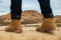 Pernas em viajante sem rosto em botas marrons e jeans azuis em pé na estrada de areia suja com montanha e céu em fundo turvo em Bardenas Reales, Navarra, Espanha — Fotografia de Stock
