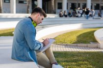 Вид збоку вдумливого студента-чоловіка в яскравих навушниках, що вивчають на університетській площі та пишуть у зошиті, що сидить на паркані з схрещеною ногою — стокове фото