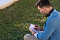 Nachdenklicher männlicher Student mit hellen Kopfhörern, der auf dem Universitätsplatz studiert und in ein Notizbuch schreibt, das am Zaun sitzt — Stockfoto