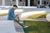 Vista lateral de estudante masculino pensativo em fones de ouvido brilhantes estudando na praça da universidade e escrevendo em livro de notas sentado em cerca com perna cruzada — Fotografia de Stock