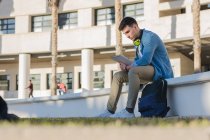 Vista lateral del estudiante masculino reflexivo en auriculares brillantes que estudian en la plaza de la universidad y escriben en un cuaderno sentado en la valla - foto de stock