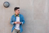 Homem elegante em fones de ouvido brilhantes surfando celular enquanto se inclina na parede de mármore em dia ensolarado olhando para longe — Fotografia de Stock