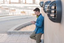 Side view of stylish man in bright headphones surfing mobile phone while leaning on marble wall in sunny day — Stock Photo
