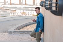 Side view of stylish man in bright headphones surfing mobile phone while leaning on marble wall in sunny day — Stock Photo