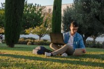 Вид збоку чоловіка з рюкзаком, який навчається в ноутбуці і пише на блокноті, сидячи в парковій траві з схрещеними ногами в сонячний день — стокове фото