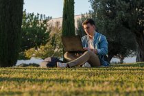 Hombre concentrado con mochila estudiando en el portátil sentado en la hierba del parque con las piernas cruzadas en el día soleado - foto de stock