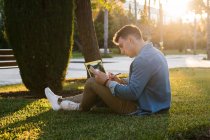 Вид збоку концентрованого чоловіка з рюкзаком, який навчається в ноутбуці і пише на блокноті, сидячи в парковій траві з схрещеними ногами в сонячний день — стокове фото