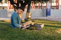 Концентрированный мужчина с рюкзаком учится за ноутбуком и пишет на блокноте, сидя в парке на траве со скрещенными ногами в солнечный день — стоковое фото