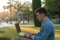 Side view of concentrated man with backpack studying at laptop sitting in park grass with crossed legs in sunny day — Stock Photo