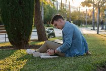 Вид сбоку сосредоточенного человека с рюкзаком, который учится на ноутбуке и пишет на блокноте, сидя в парке на траве со скрещенными ногами в солнечный день — стоковое фото
