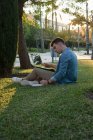 Vista lateral del hombre con mochila estudiando en el portátil y escribiendo en un bloc de notas sentado en el césped del parque con las piernas cruzadas en el día soleado - foto de stock