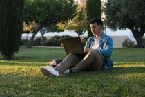 Konzentrierter Mann mit Rucksack am Laptop sitzend im Parkgras mit überkreuzten Beinen bei sonnigem Tag — Stockfoto