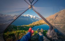 Cultivez d'agréables randonneurs en baskets colorées allongées avec des jambes croisées dans une petite tente transparente près d'un lac clair en montagne par temps ensoleillé à Chamonix, Mont-Blanc — Photo de stock