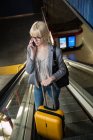 Young blonde businesswoman riding on moving stairs with luggage and talking on smartphone — Stock Photo