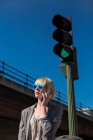 Блондинка в солнечных очках разговаривает на смартфоне и смотрит в сторону зеленого светофора на улице — стоковое фото