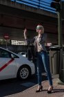 Молодая стильная деловая женщина жестикулирует для остановки такси и разговаривает на смартфоне на городской улице — стоковое фото