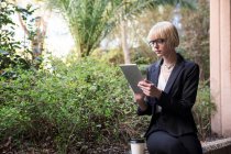 Jovem mulher de negócios loira sentada com copo de papel e usando tablet digital no jardim — Fotografia de Stock