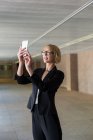Blonde businesswoman in formal clothes taking selfie or browsing smartphone in big hall — Stock Photo