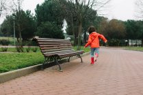Visão traseira da criança alegre adorável anônima em capa de chuva vermelha e botas de borracha se divertindo correndo na rua no parque em dia cinza — Fotografia de Stock