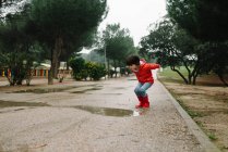 Очаровательный радостный ребенок в красном плаще и резиновых сапогах с удовольствием прыгает в луже на улице в парке в серый день — стоковое фото