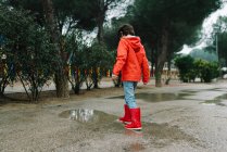 Visão traseira da criança alegre adorável anônima em capa de chuva vermelha e botas de borracha se divertindo brincando com a poça na rua no parque em dia cinza — Fotografia de Stock