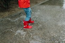 Liebenswertes fröhliches Kind in rotem Regenmantel und Gummistiefeln, das an grauen Tagen Spaß beim Springen in einer Pfütze auf der Straße im Park hat — Stockfoto