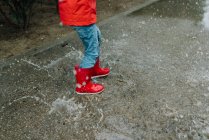 Adorable joyful child in red raincoat and rubber boots having fun jumping in puddle on street in park in gray day — Stock Photo