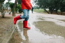 Adorabile bambino gioioso in impermeabile rosso e stivali di gomma divertirsi saltando in pozzanghera sulla strada in parco in giornata grigia — Foto stock