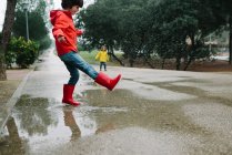 Adoráveis crianças alegres em capa de chuva vermelha e amarela e botas de borracha se divertindo jogando na poça na rua no parque em dia cinza — Fotografia de Stock