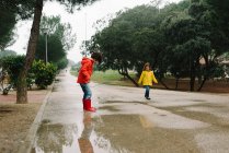 Adorable joyful children in red and yellow raincoat and rubber boots having fun playing in puddle in street in park in gray day — Stock Photo